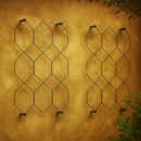 Twin geometric metal trellises mounted on a textured mustard yellow wall with soft natural lighting and greenery in the foreground.