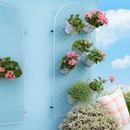 Wall-mounted white trellises with potted pink flowers on a light blue wall, under a sunny sky.